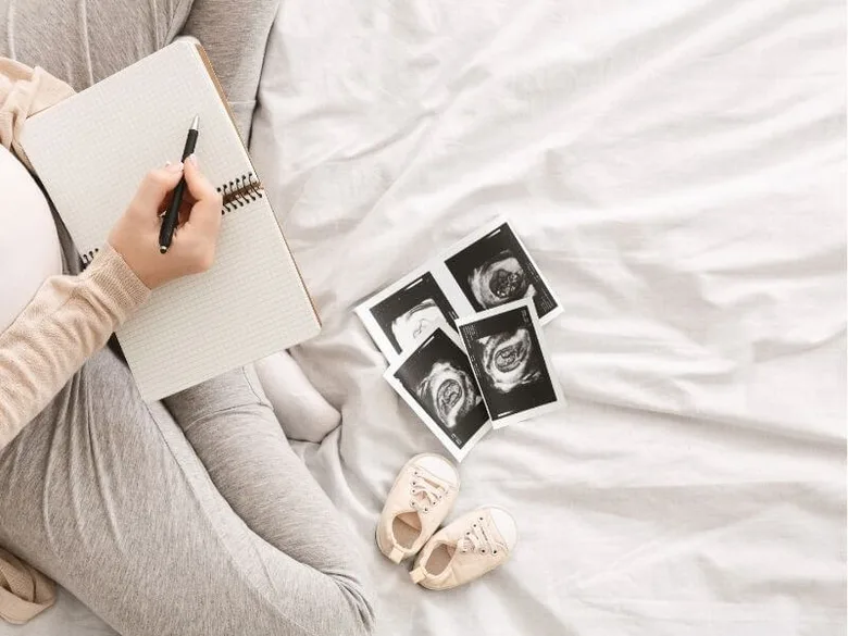Těhotná žena píše do sešitu vedle ultrazvukových snímků a dětských botiček.